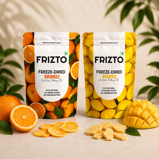 Two stand-up pouches of Frizto freeze-dried fruit cracker are displayed on a light beige surface with fresh fruit props. The left pouch is labeled 'FRIZTO FREEZE-DRIED ORANGE' and features orange imagery, while the right pouch is labeled 'FRIZTO FREEZE-DRIED MANGO' and features yellow mango imagery.