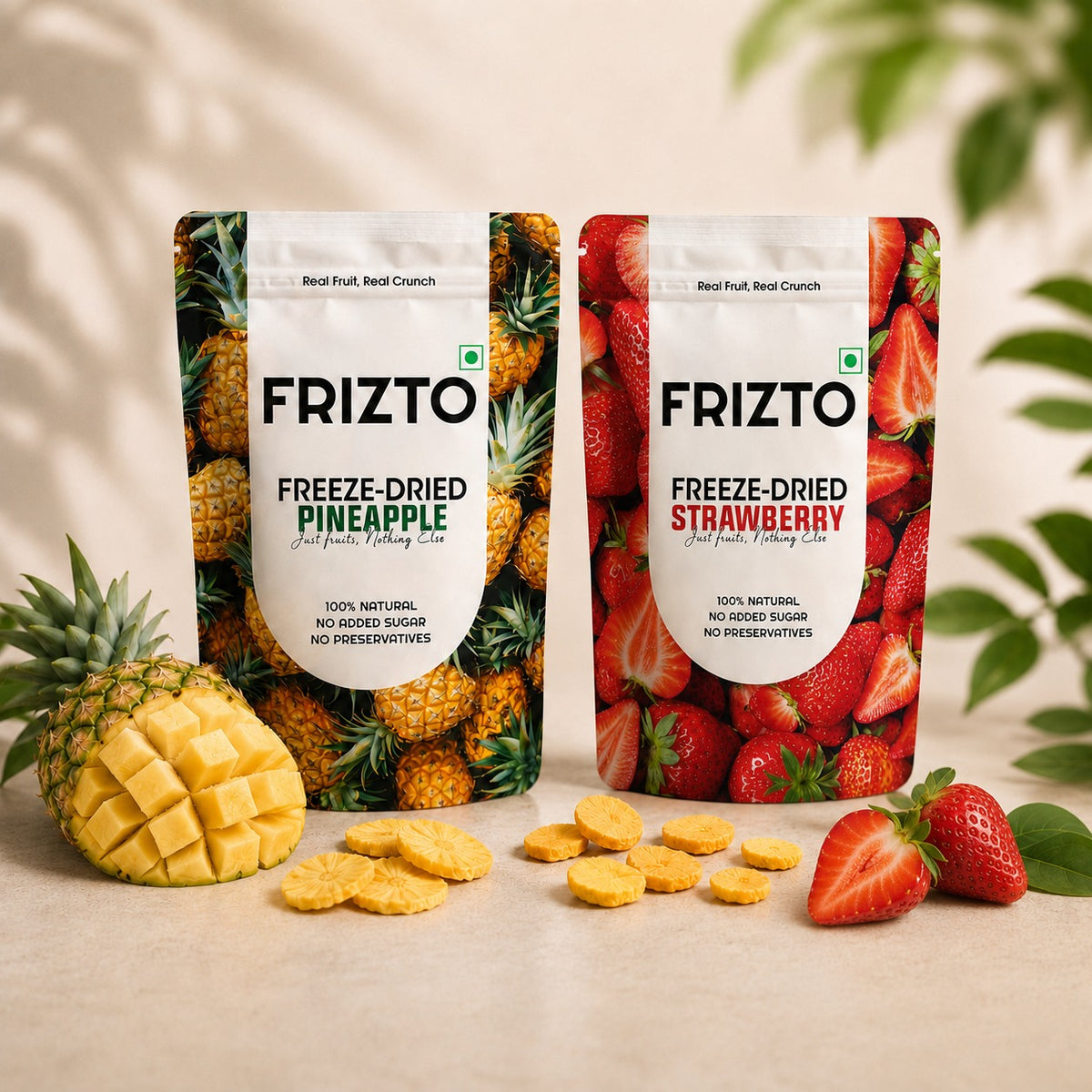 Two FRIZTO Freeze-Dried Fruit Snacks pouches, Pineapple and Strawberry, displayed on a light beige surface with fresh pineapple, sliced pineapple, strawberries, and scattered yellow snack pieces. The background is a warm beige with soft leaf shadows.