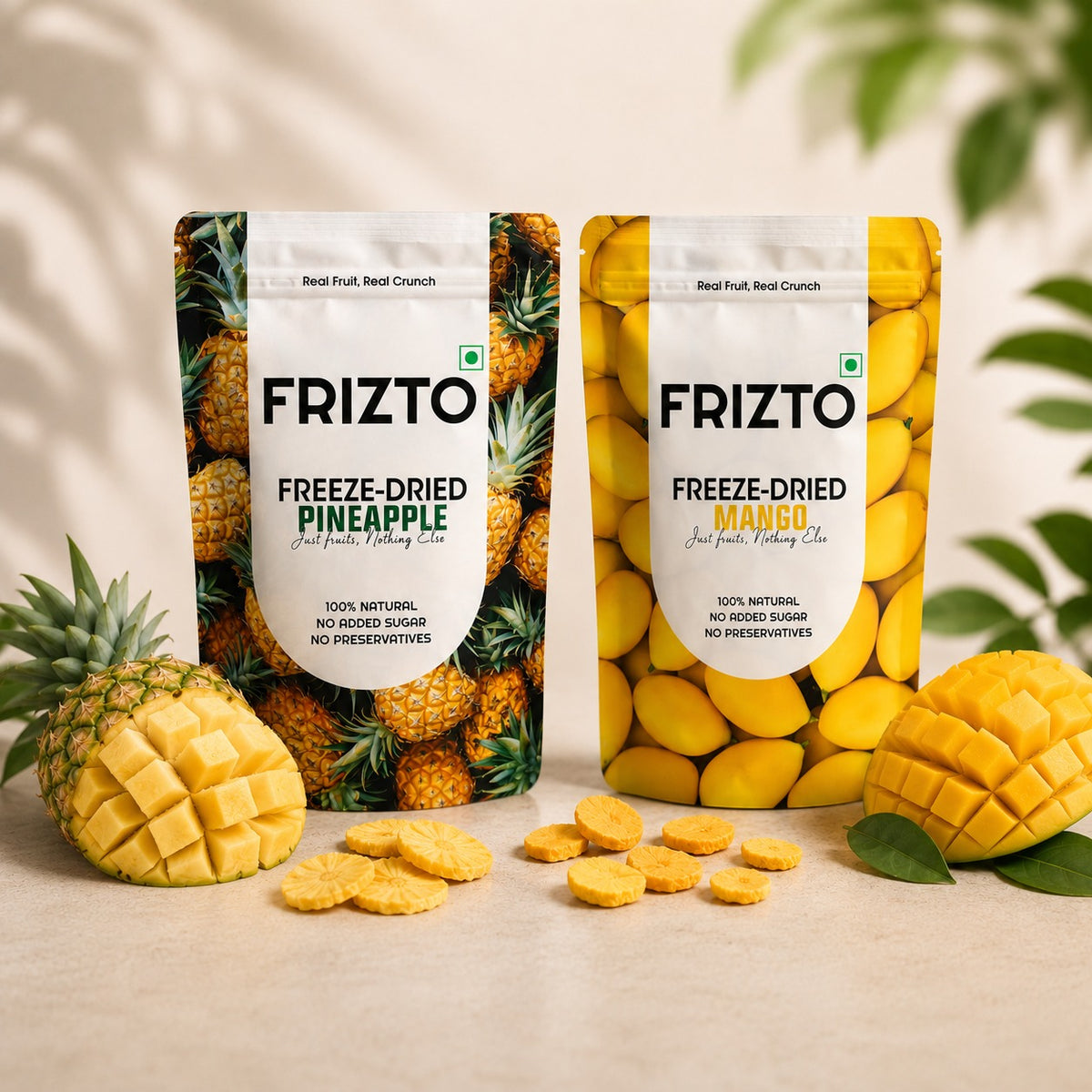 Two FRIZTO freeze-dried fruit punch bags, one pineapple flavor and one mango flavor, displayed on a light beige surface with fresh pineapple, mango, and scattered snack pieces. The background is softly blurred with green leaves.