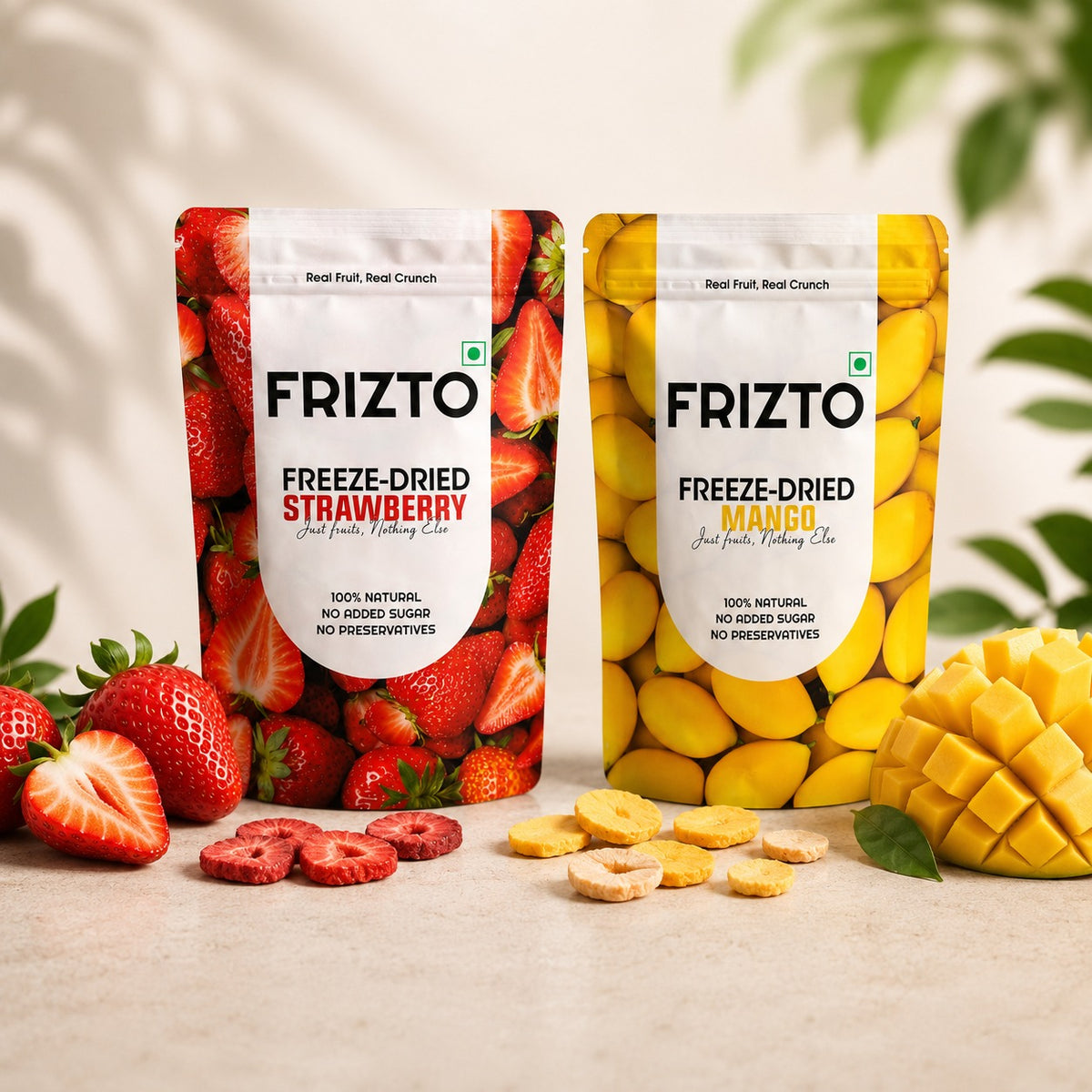 Two stand-up pouches of FRIZTO freeze-dried fruit crunch are shown on a light surface with fresh strawberries and mango slices around them. The left pouch is strawberry flavor with red fruit imagery, and the right pouch is mango flavor with yellow fruit imagery.